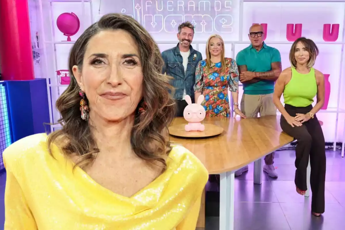 Un grupo de personas posando en un set de televisión colorido, con una mujer en primer plano usando un vestido amarillo brillante y otras cuatro personas de pie y sentadas alrededor de una mesa con un muñeco rosa en el centro.