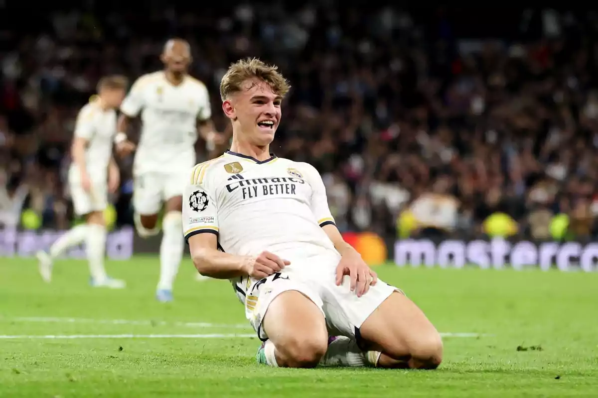 Jugador de fútbol celebrando un gol en el campo con compañeros de equipo al fondo.