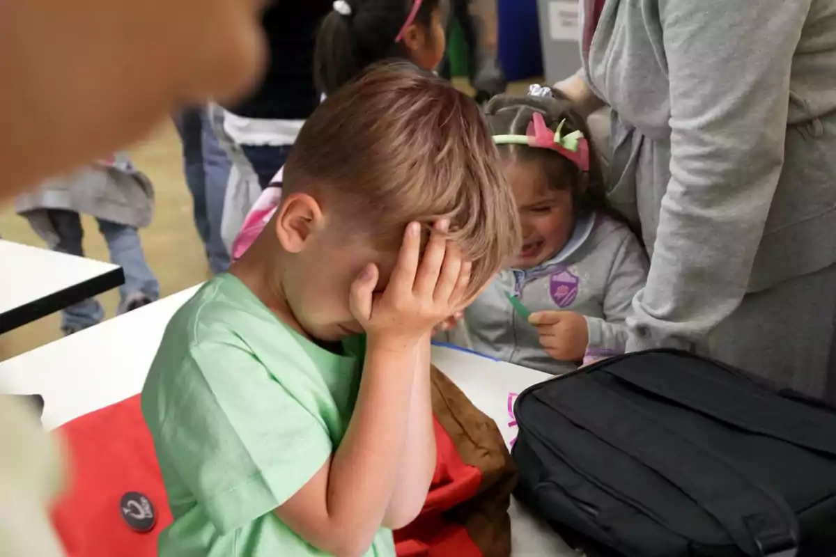 Niño con camiseta verde cubriéndose el rostro con la mano mientras una niña llora a su lado en un ambiente escolar