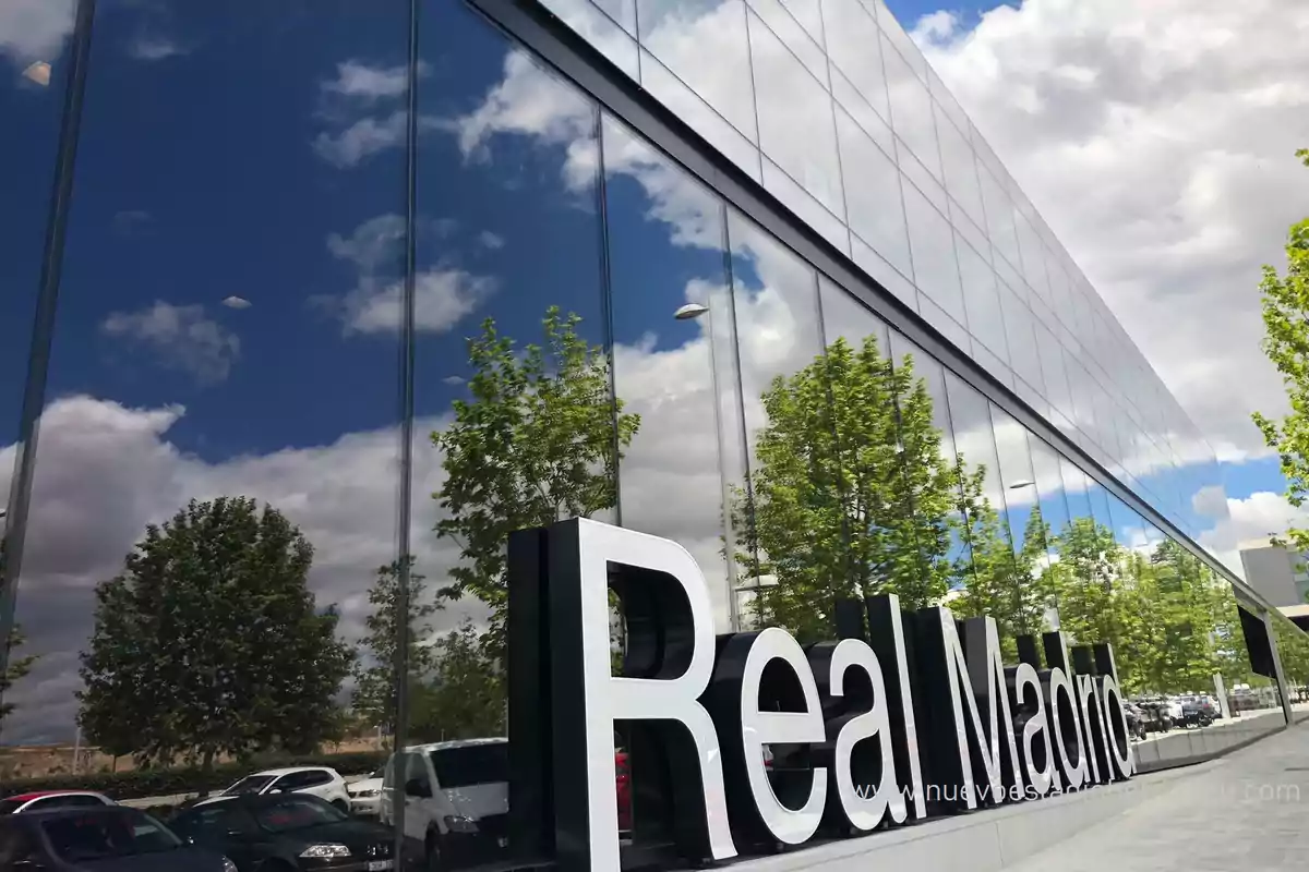 Edificio moderno con fachada de vidrio reflejando el cielo y árboles, con un letrero grande que dice "Real Madrid" en la parte inferior derecha.