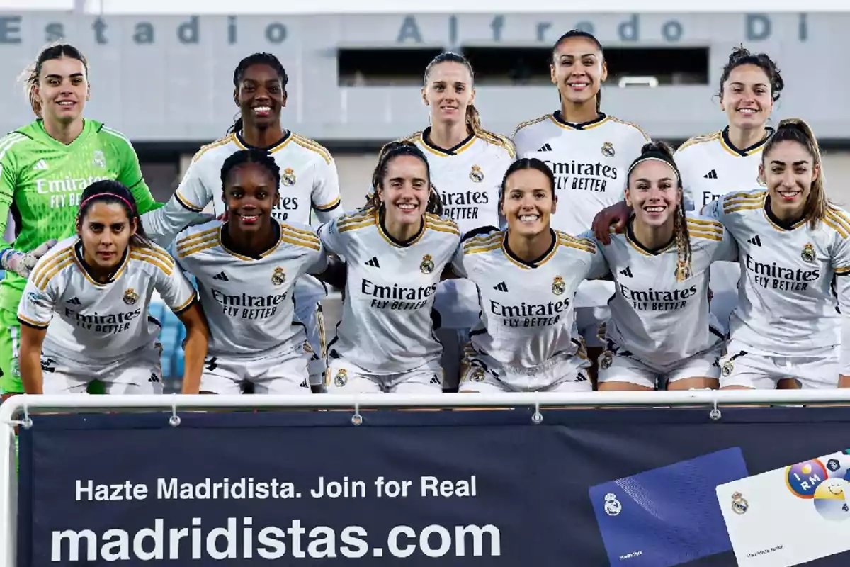 Un equipo de fútbol femenino posando para una foto grupal en el estadio Alfredo Di Stéfano, con camisetas blancas del Real Madrid y un cartel promocional de madridistas.com al frente.