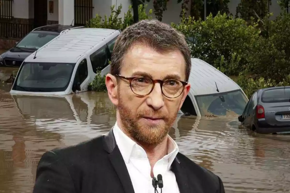 Un hombre con gafas y barba en primer plano con una escena de inundación y coches sumergidos en el fondo.