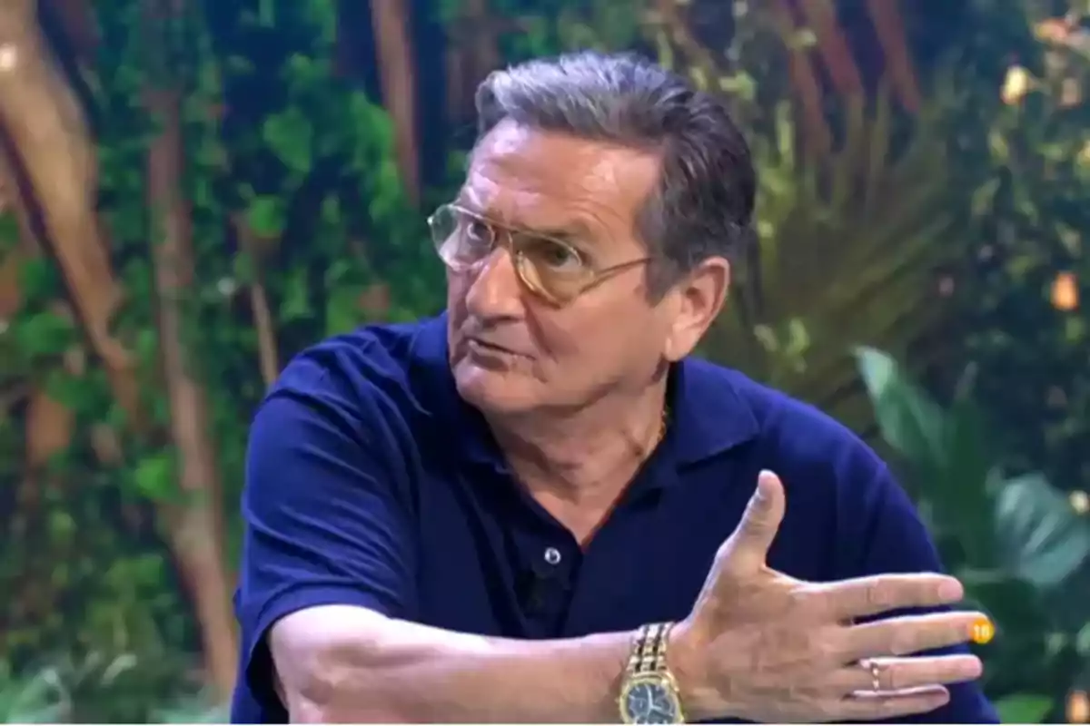 Un hombre con gafas y reloj dorado gesticula mientras habla en un entorno con vegetación de fondo.