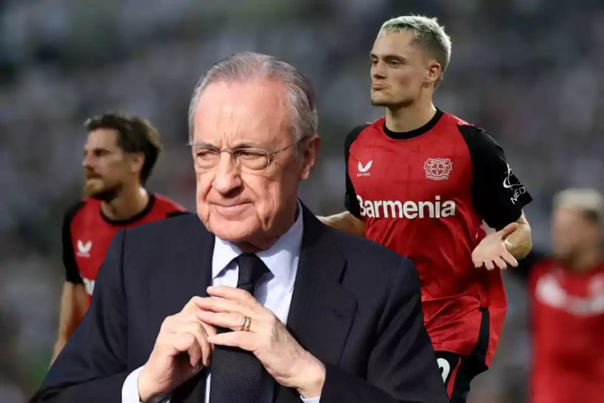 Un hombre mayor con traje y gafas está en primer plano mientras dos jugadores de fútbol con camisetas rojas están en el fondo.