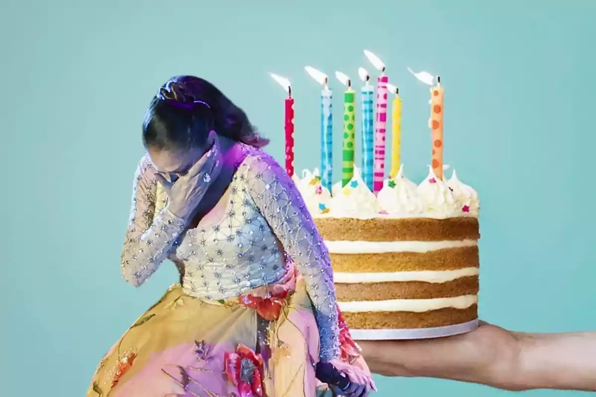 Una mujer con un vestido elegante se cubre la cara con una mano mientras una mano sostiene un pastel de cumpleaños con velas encendidas en el fondo.