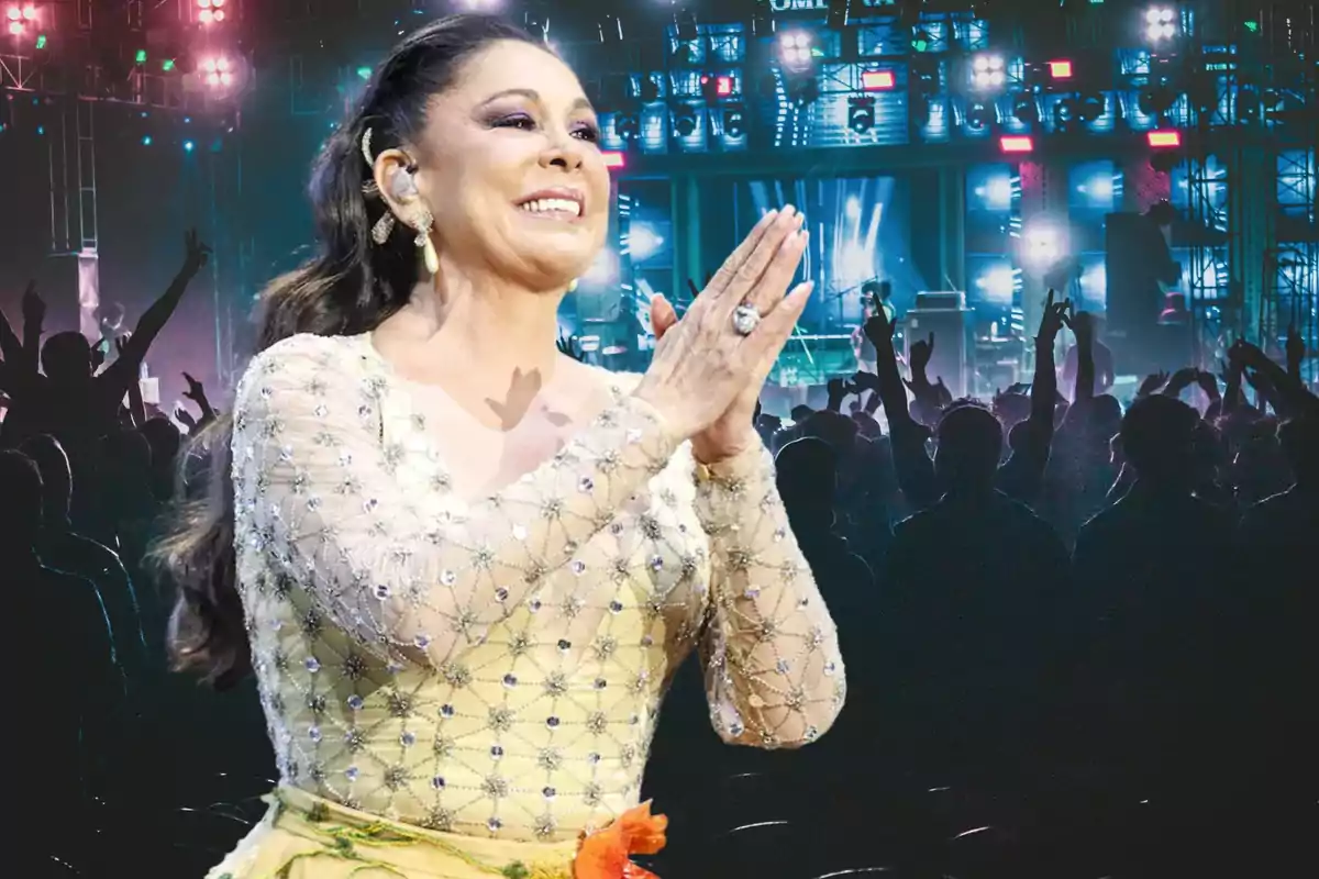 Una mujer sonriente con un vestido elegante aplaude en un escenario iluminado, mientras una multitud de personas la observa y celebra en un concierto.