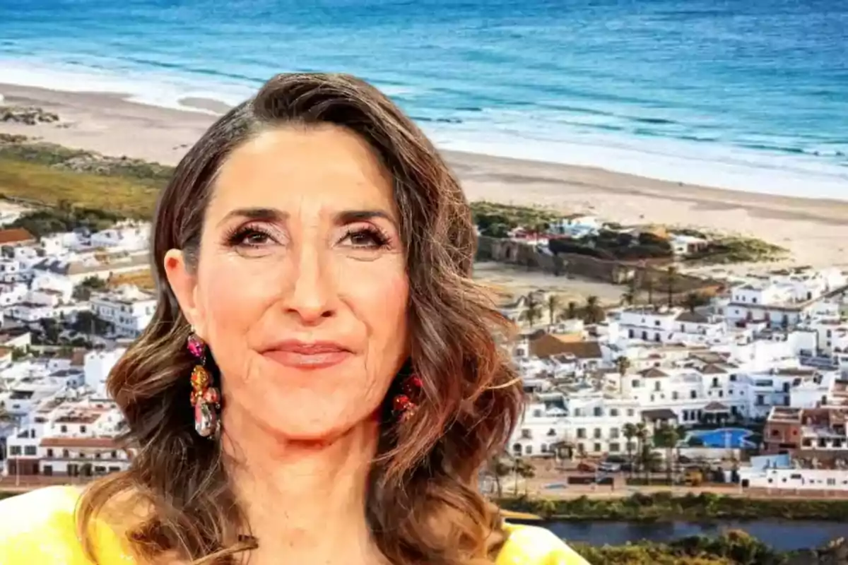 Mujer de cabello castaño y ondulado con pendientes coloridos posando frente a un pueblo costero con casas blancas y el mar de fondo