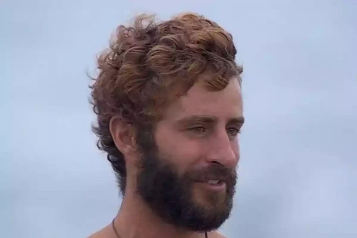 Un hombre con barba y cabello rizado mirando hacia un lado con un fondo de cielo nublado. Un hombre con barba y cabello rizado mirando hacia un lado con un fondo de cielo nublado.