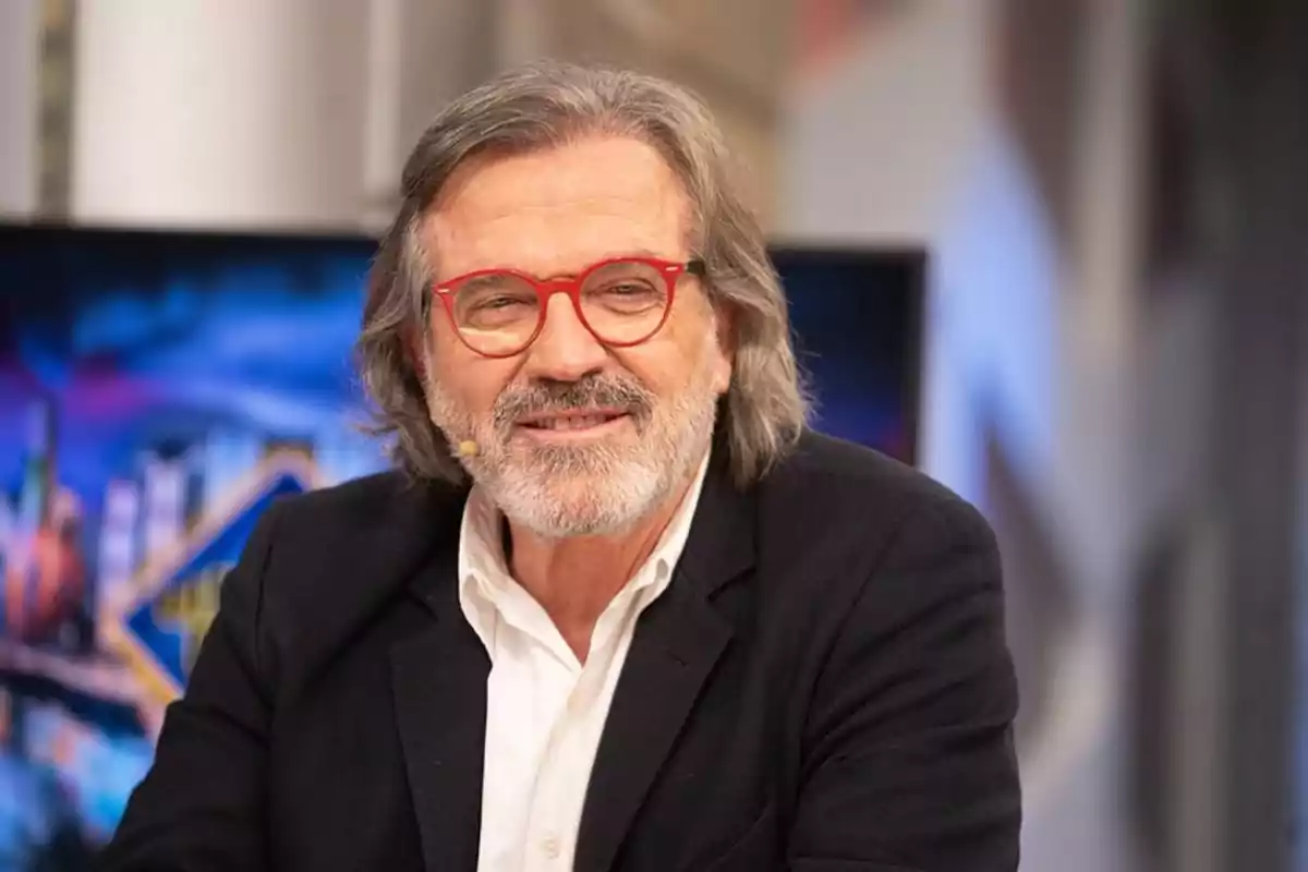 Un hombre con gafas rojas y barba gris sonríe mientras está sentado en un estudio de televisión. Un hombre con gafas rojas y barba gris sonríe mientras está sentado en un estudio de televisión.