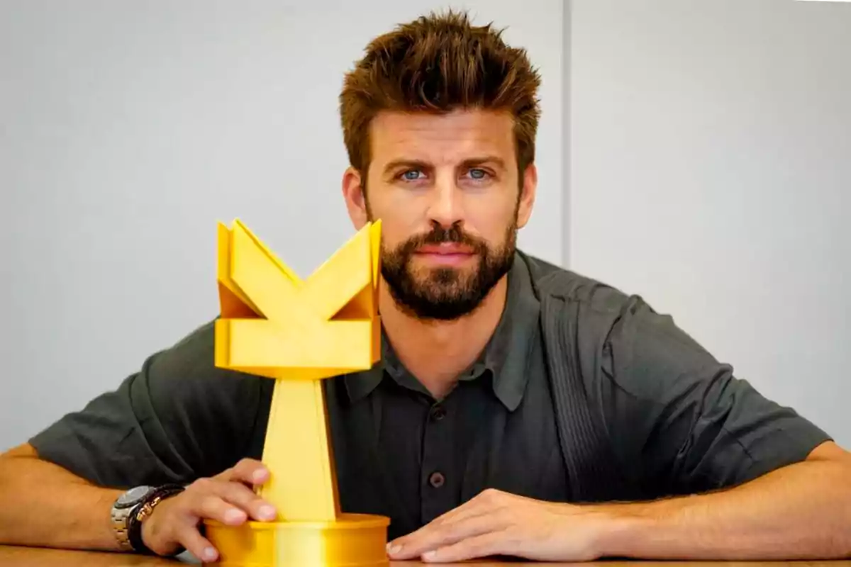 Un hombre con barba y cabello castaño sostiene un trofeo dorado en forma de Y sobre una mesa.