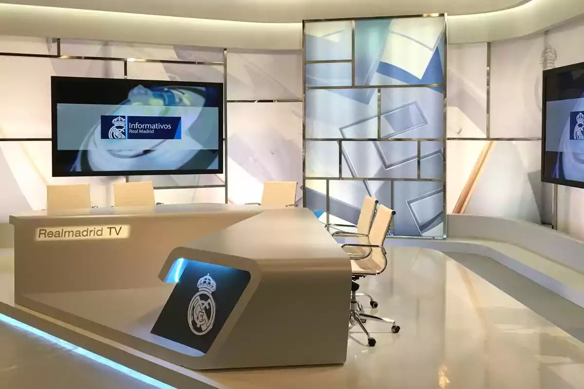 Estudio de televisión de Real Madrid TV con un set moderno y elegante, sillas blancas y una pantalla grande mostrando "Informativos Real Madrid".