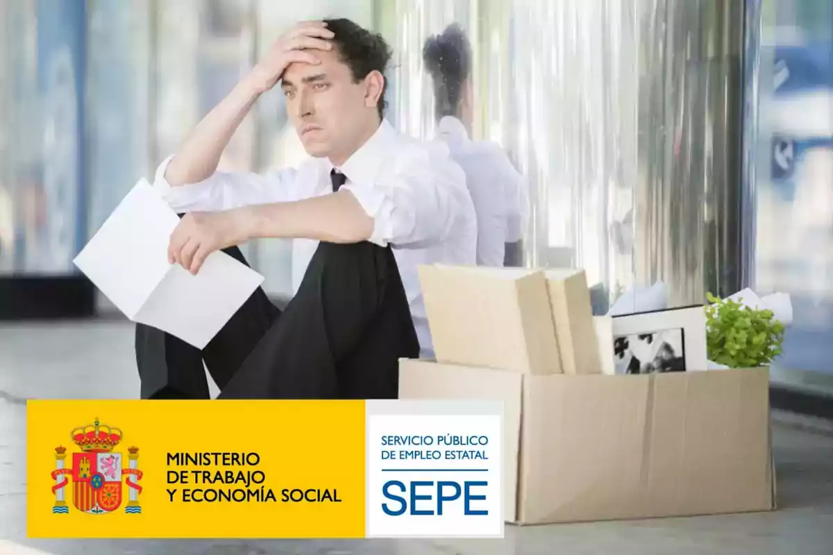 Un hombre con expresión preocupada está sentado en el suelo junto a una caja de cartón con objetos personales, sosteniendo un papel en la mano. En la parte inferior de la imagen, se encuentran los logotipos del Ministerio de Trabajo y Economía Social y del Servicio Público de Empleo Estatal (SEPE).