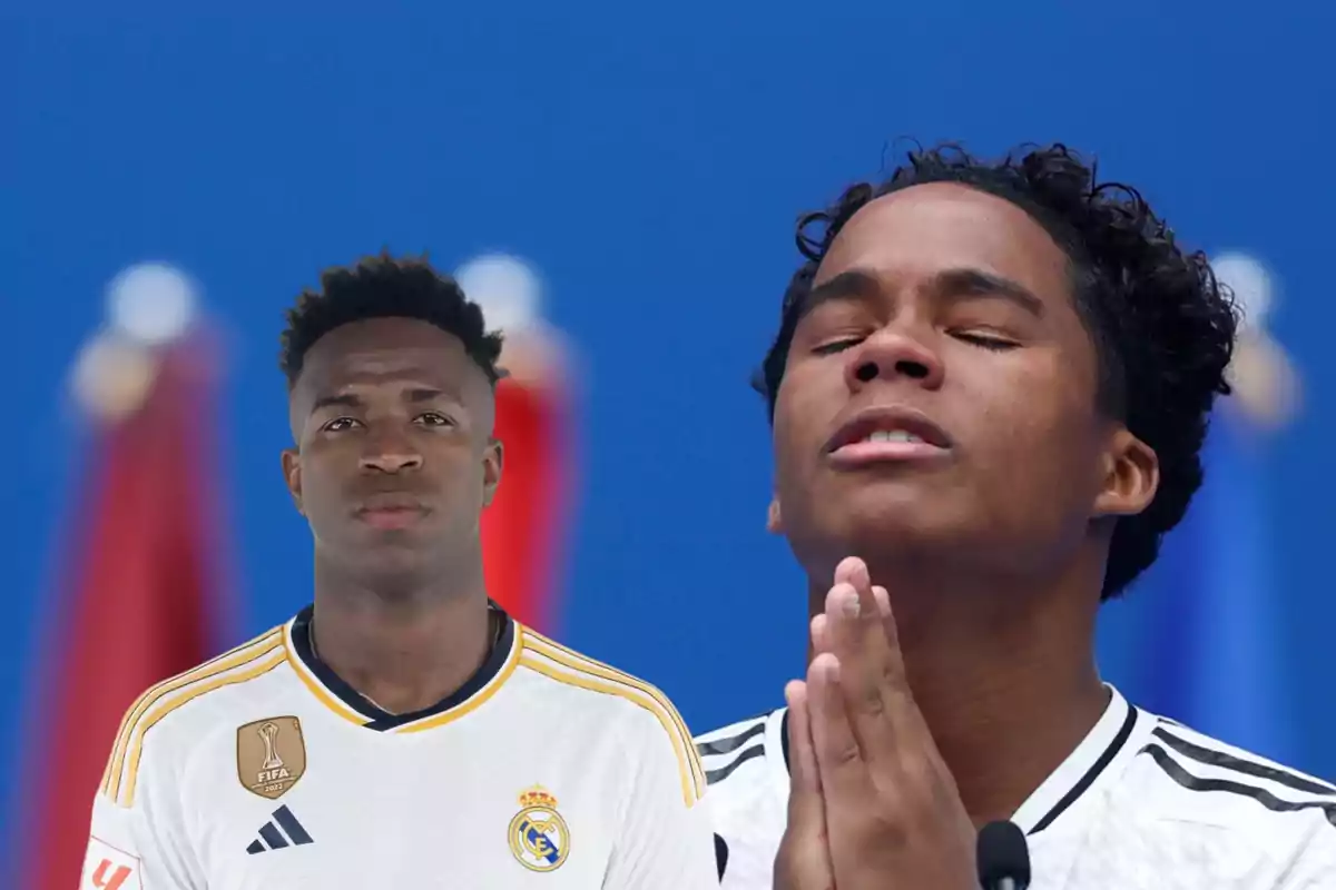 Dos jugadores de fútbol con camisetas blancas, uno con los ojos cerrados y las manos juntas en actitud de oración y el otro mirando al frente con expresión seria, con un fondo azul y banderas desenfocadas.