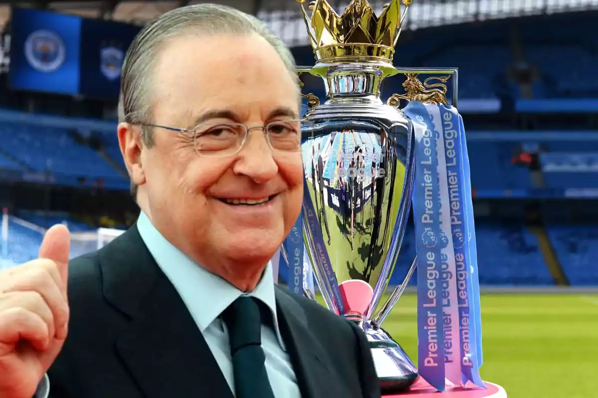 Un hombre sonriente con gafas y traje oscuro está junto a un trofeo de la Premier League en un estadio de fútbol.