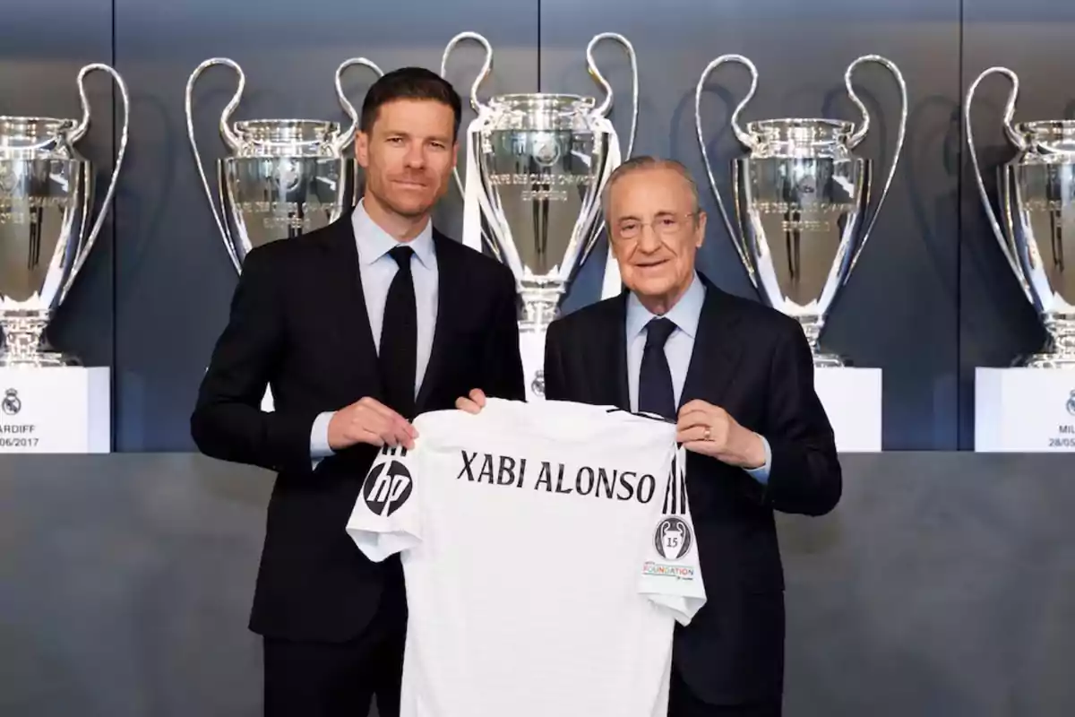 Dos hombres con traje sostienen una camiseta de fútbol con el nombre Xabi Alonso frente a varios trofeos grandes de fondo Dos hombres con traje sostienen una camiseta de fútbol con el nombre Xabi Alonso frente a varios trofeos grandes de fondo