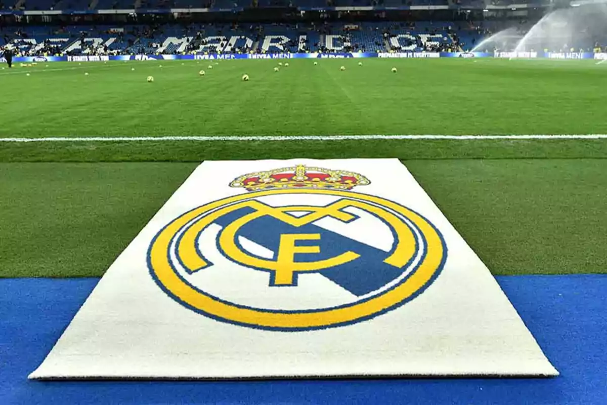 Campo de fútbol con el escudo del Real Madrid en primer plano y las gradas del estadio al fondo.