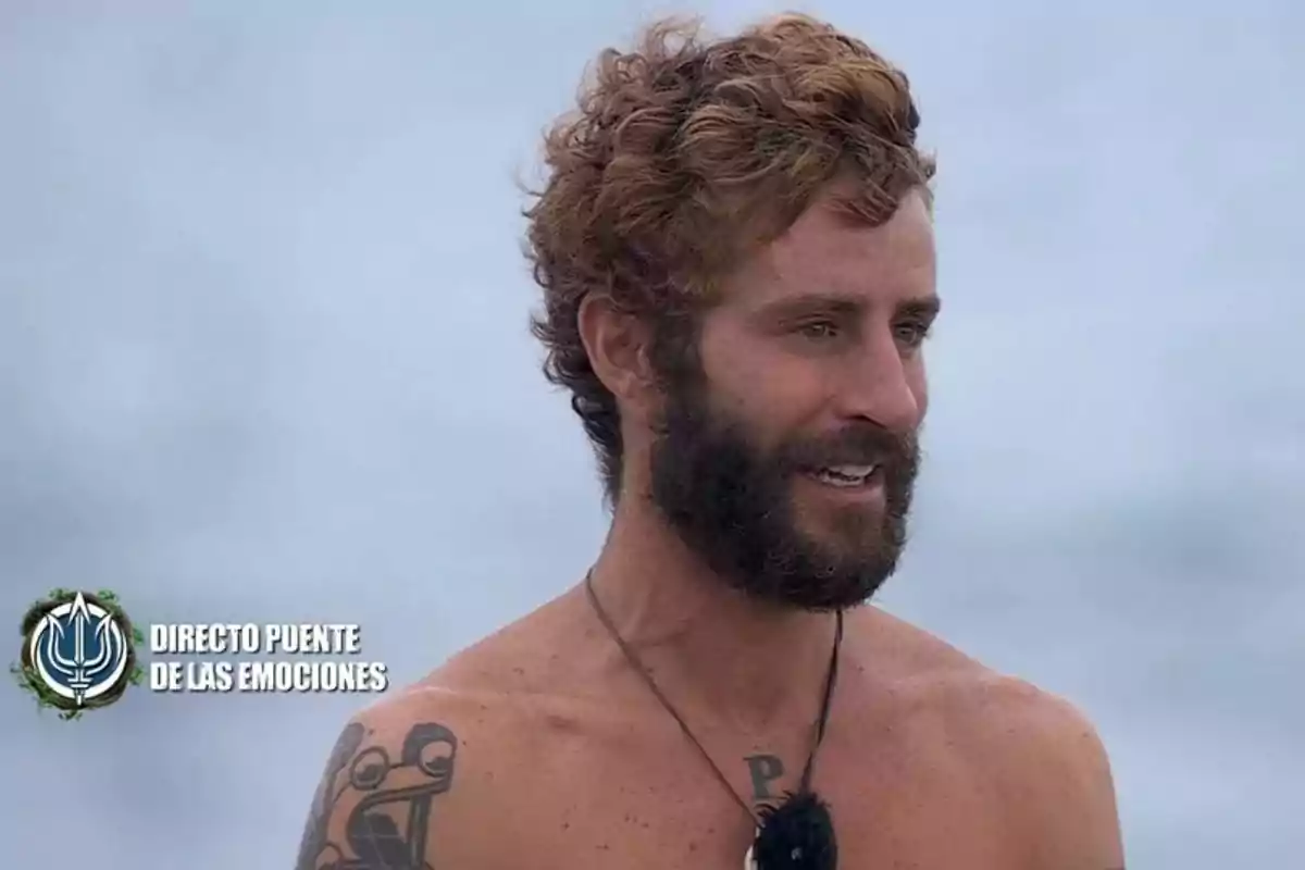 Un hombre con barba y cabello rizado está al aire libre, lleva un collar y tiene un tatuaje en el hombro, en la esquina inferior izquierda se ve un logotipo con el texto Un hombre con barba y cabello rizado está al aire libre, lleva un collar y tiene un tatuaje en el hombro, en la esquina inferior izquierda se ve un logotipo con el texto
