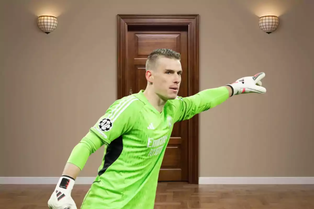 Un portero de fútbol con uniforme verde señala con su mano derecha en una habitación con una puerta de madera y dos lámparas de pared.