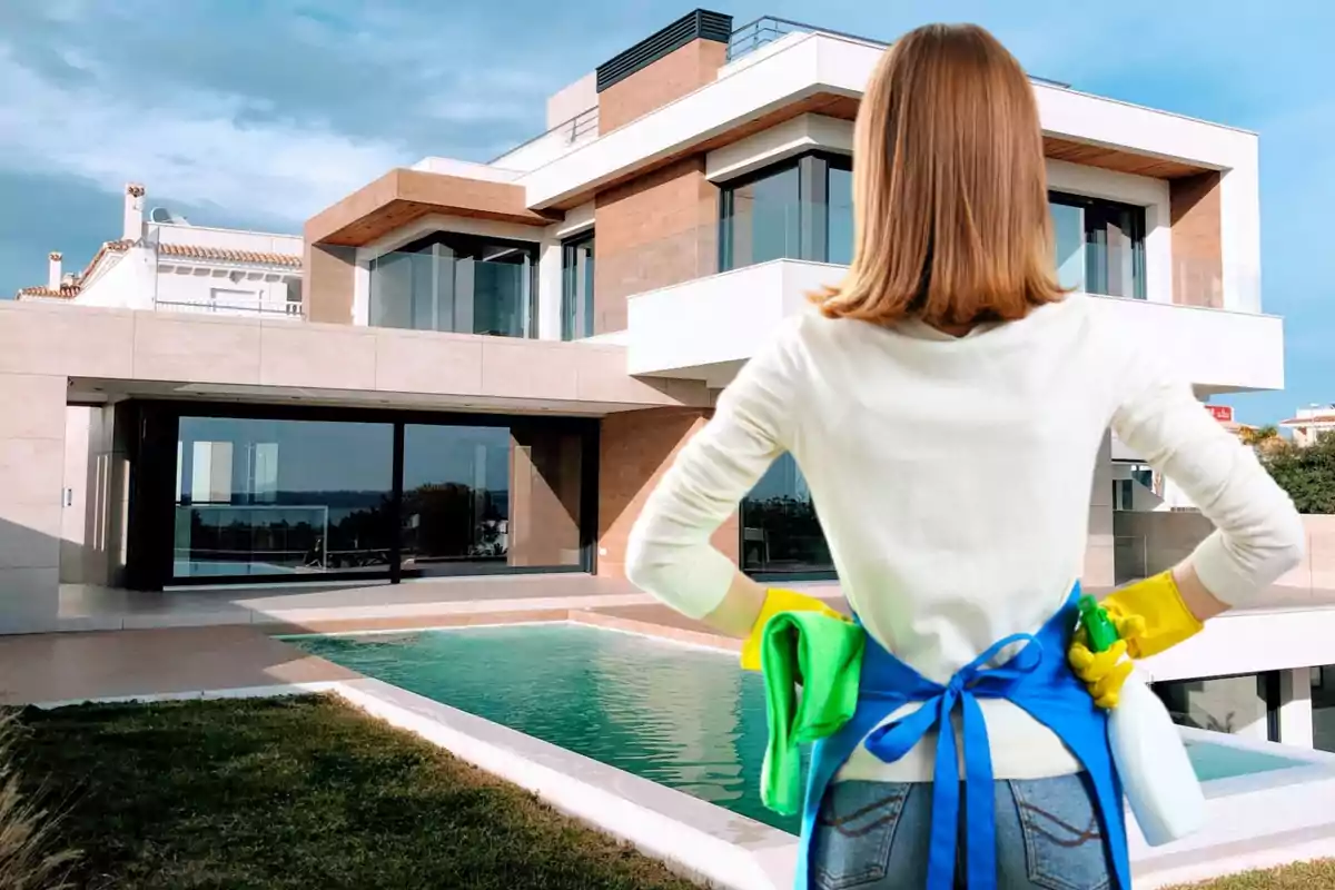 Persona con guantes de limpieza y delantal azul mirando una casa moderna con piscina.