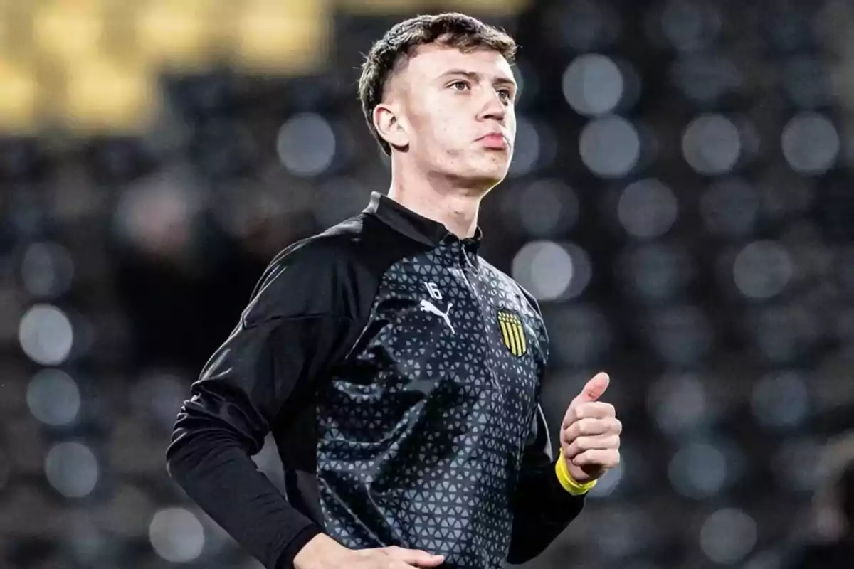 Un jugador de fútbol con uniforme de entrenamiento negro y gris, con el logo de Puma y un escudo amarillo en el pecho, corriendo en un estadio con luces desenfocadas al fondo.