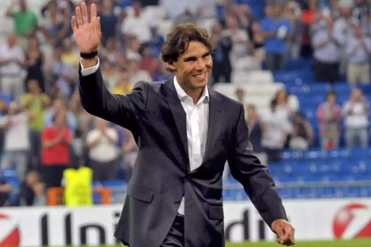 Un hombre sonriente con traje oscuro saluda con la mano levantada en un estadio lleno de gente.