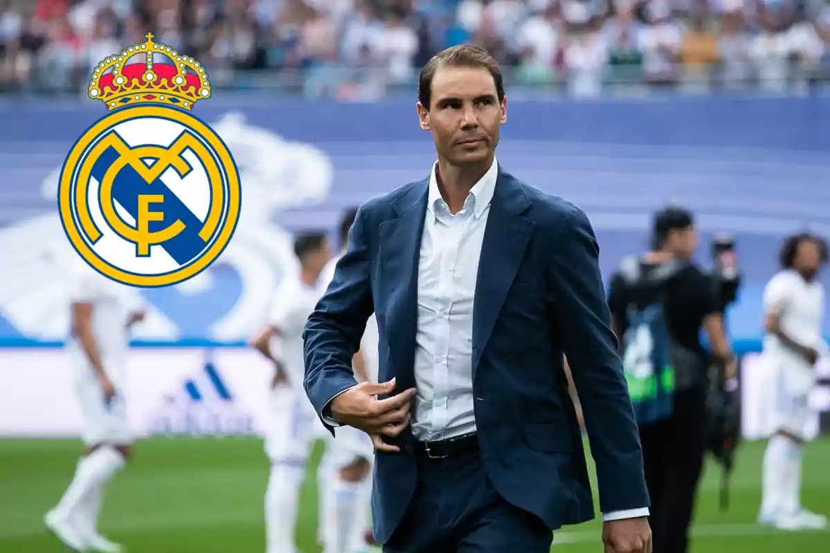 Un hombre con traje azul camina en un estadio de fútbol con el logo del Real Madrid al fondo.