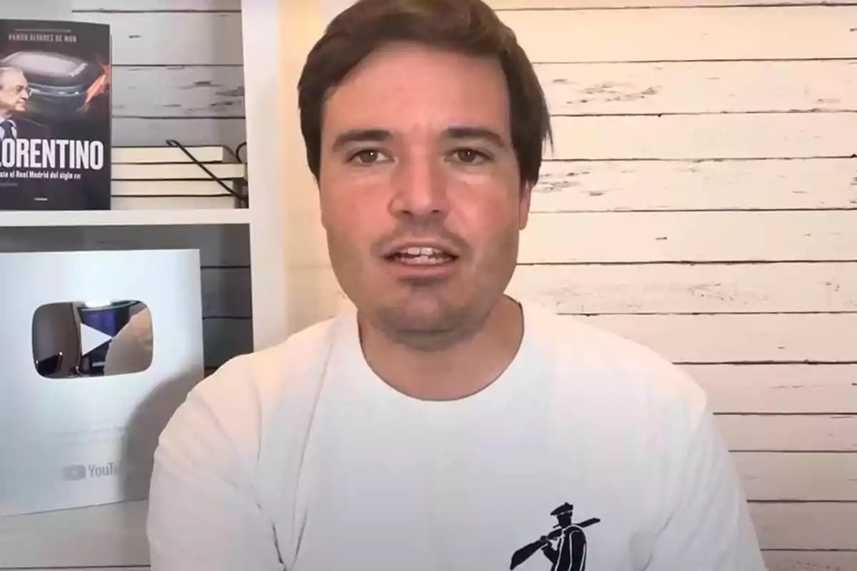 Un hombre con una camiseta blanca está sentado frente a una estantería que contiene un libro y una placa de YouTube.