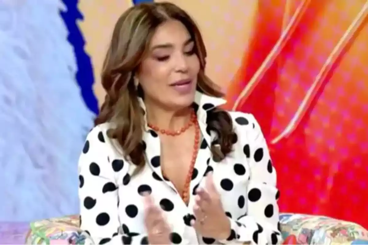 Mujer con blusa de lunares negros y collar naranja gesticulando mientras está sentada en un set colorido. Mujer con blusa de lunares negros y collar naranja gesticulando mientras está sentada en un set colorido.