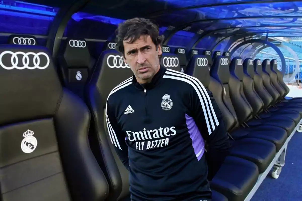 Un hombre con uniforme de entrenamiento del Real Madrid está de pie frente a un banquillo vacío en un estadio.