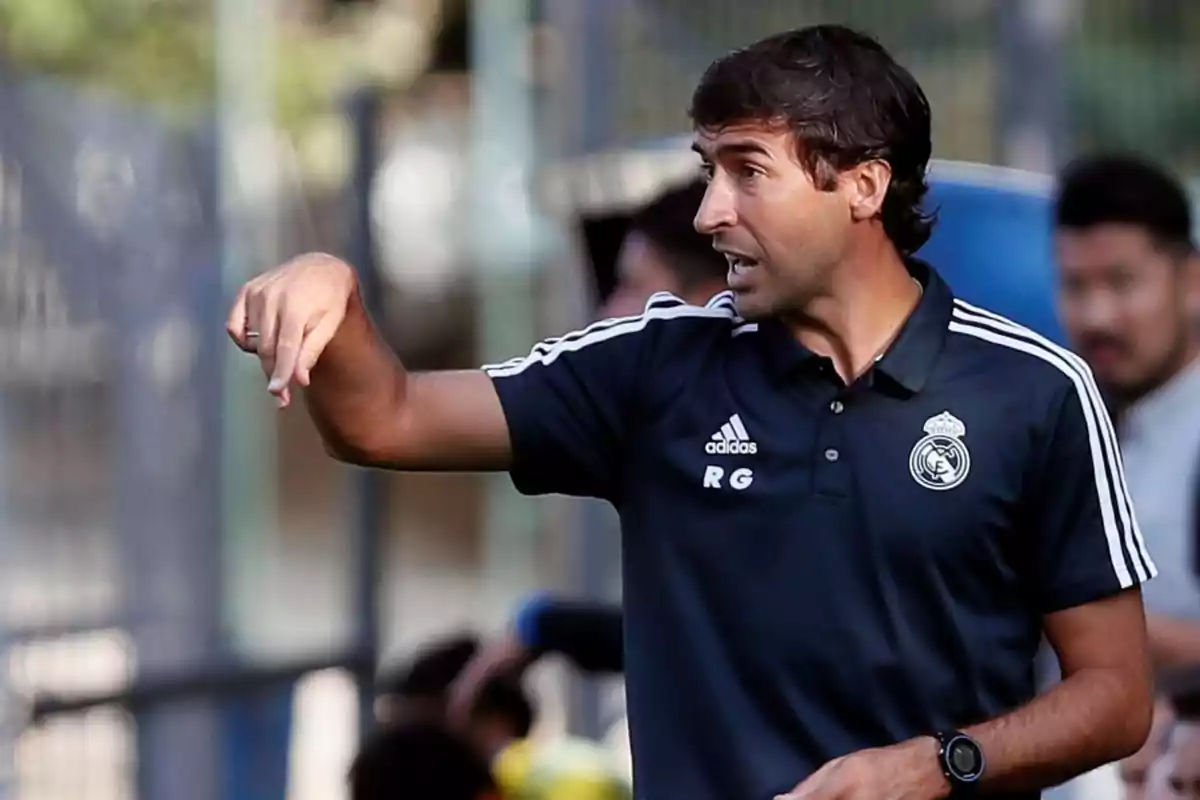 Un entrenador de fútbol con uniforme de entrenamiento del Real Madrid gesticula mientras da instrucciones en un campo de entrenamiento.