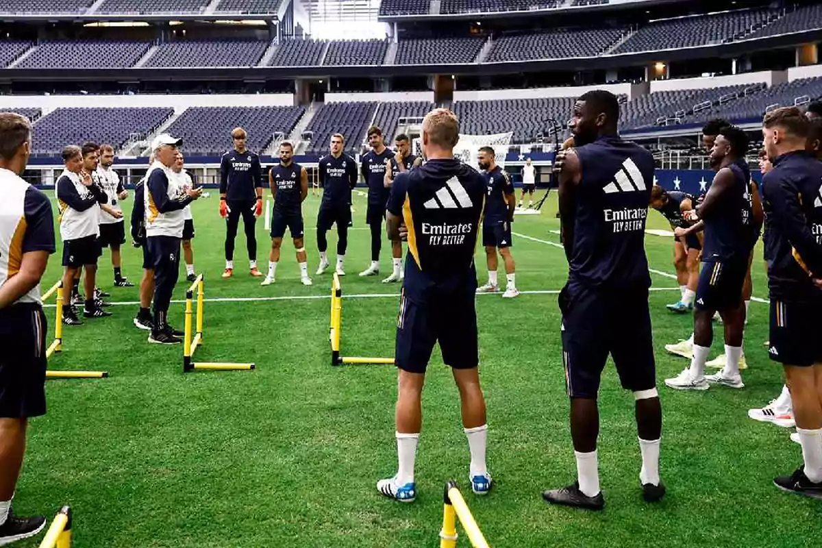 Un grupo de jugadores de fútbol y entrenadores se encuentran en un campo de entrenamiento dentro de un estadio, escuchando instrucciones mientras están rodeados de equipo de entrenamiento.