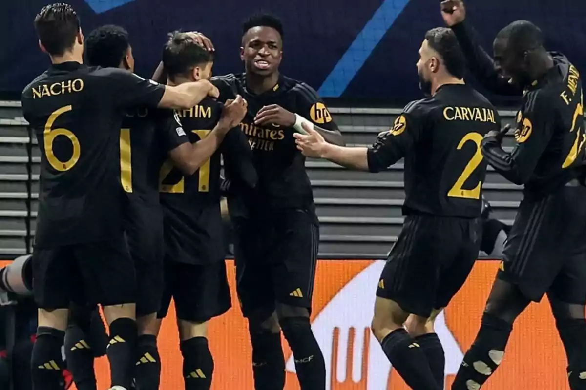 Jugadores de fútbol celebrando un gol en el campo, vistiendo uniformes negros con detalles dorados y números visibles en la espalda.