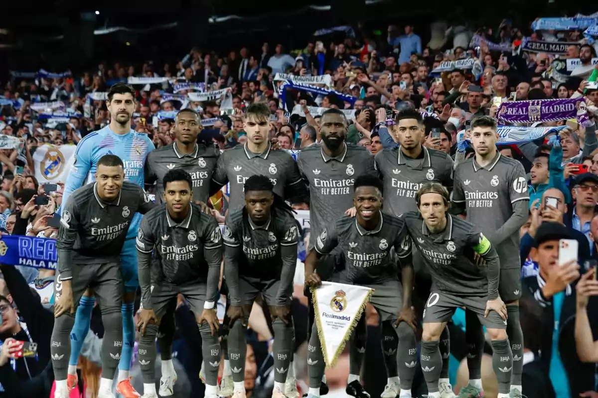 Un equipo de fútbol posando para una foto grupal en el campo con jugadores vistiendo uniformes oscuros y un portero con uniforme azul claro mientras los aficionados sostienen bufandas en el fondo.