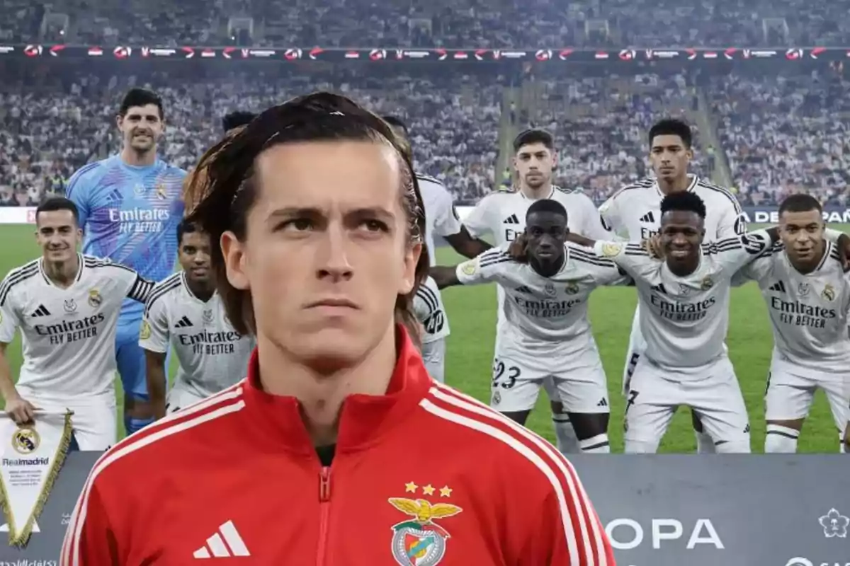 Un jugador con uniforme rojo del Benfica está en primer plano mientras un equipo de fútbol con uniformes blancos posa para una foto grupal en el fondo en un estadio lleno de espectadores.