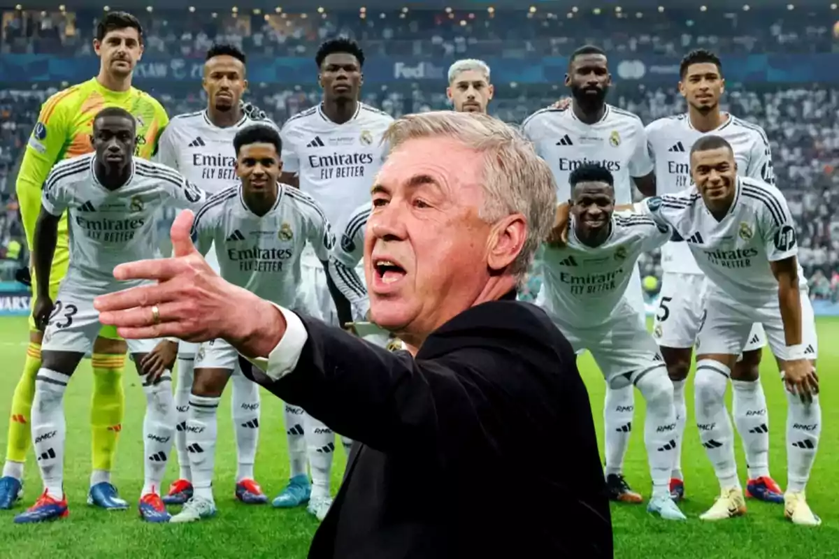 Un entrenador de fútbol gesticula frente a un equipo de jugadores en uniforme blanco posando en un estadio.