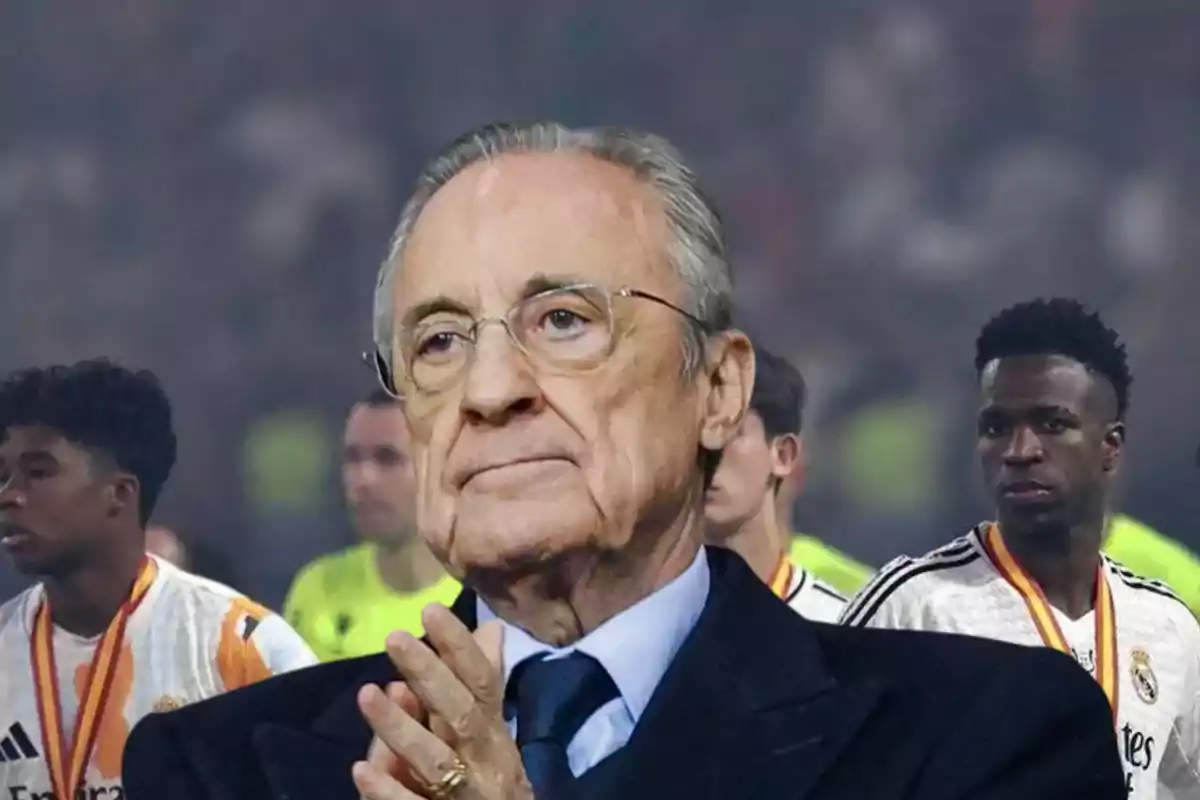 Un hombre mayor con gafas y traje oscuro aplaude mientras varios jugadores de fútbol con medallas están en el fondo.