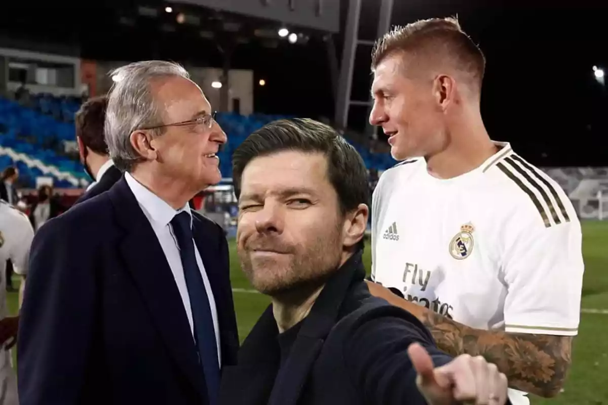 Tres hombres en un estadio, uno de ellos con traje y otro con uniforme de fútbol conversan mientras una tercera persona en primer plano hace un gesto de selfie guiñando un ojo.