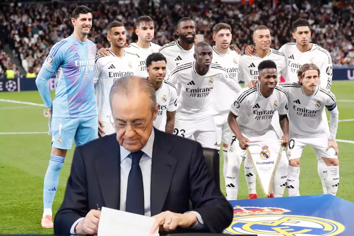 Un grupo de jugadores de fútbol con uniforme blanco posan en un estadio lleno de espectadores mientras un hombre mayor, vestido con traje, está sentado frente a ellos escribiendo en un papel.