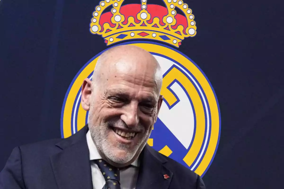 Un hombre calvo y sonriente con barba vestido con traje y corbata frente a un escudo de un equipo de fútbol.
