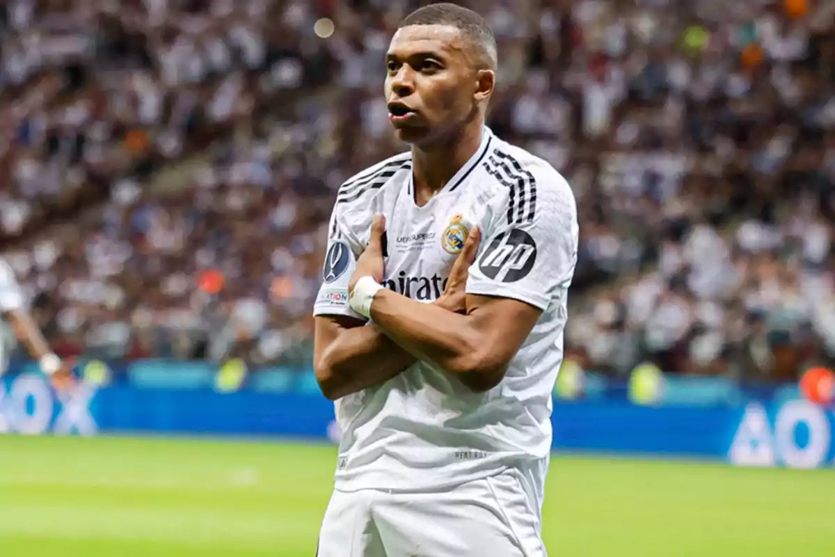 Jugador de fútbol celebrando un gol con los brazos cruzados sobre el pecho, vistiendo el uniforme blanco de su equipo en un estadio lleno de espectadores.