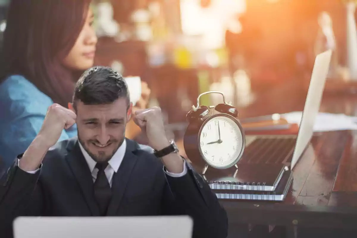 Hombre en traje celebrando frente a una computadora portátil con un reloj despertador sobre la mesa.