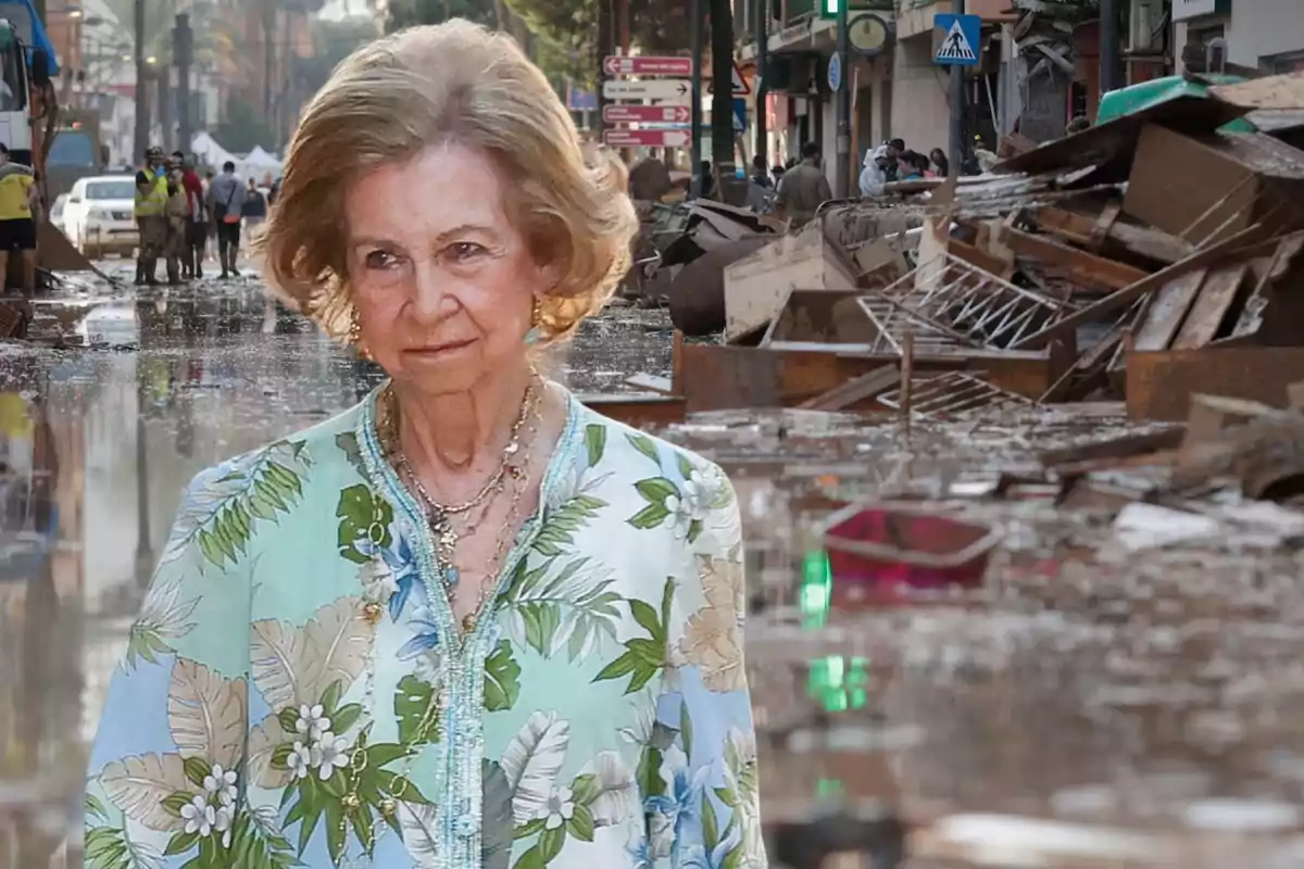 Una mujer mayor con una blusa floral está de pie en una calle inundada y llena de escombros, mientras varias personas trabajan en el fondo.