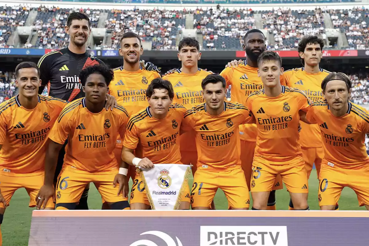 Jugadores del Real Madrid posando para una foto de equipo en el campo de fútbol, vistiendo uniformes naranjas con el logo de "Emirates Fly Better".