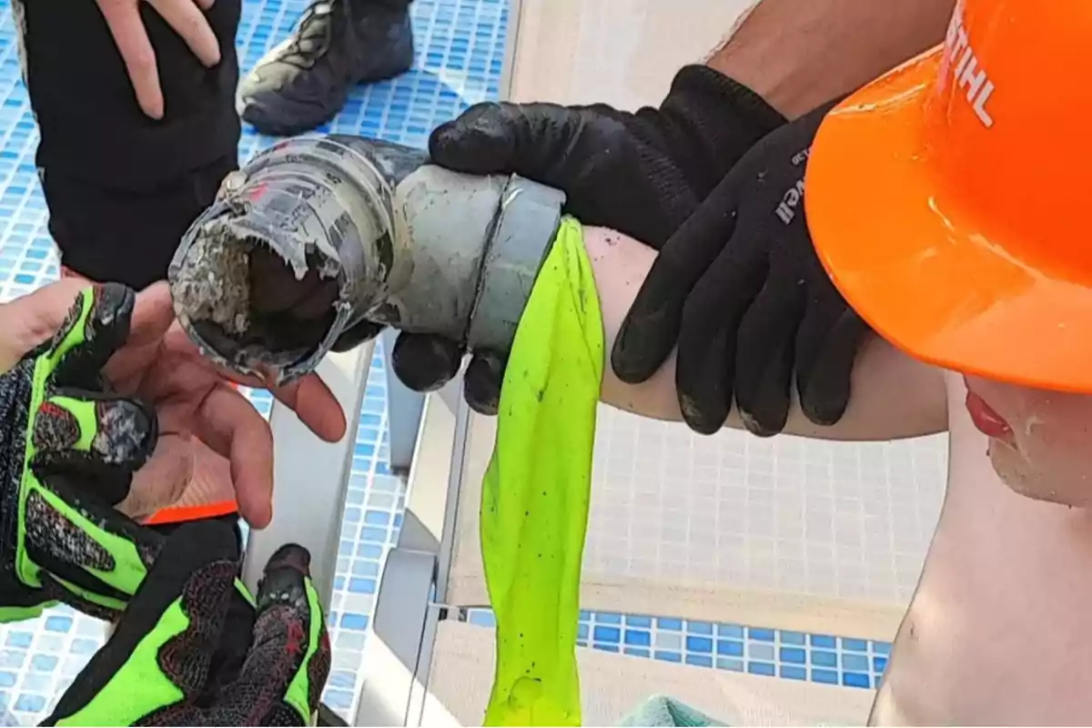 Un niño con casco naranja está siendo asistido por rescatistas que intentan liberar su brazo atrapado en una tubería junto a una piscina