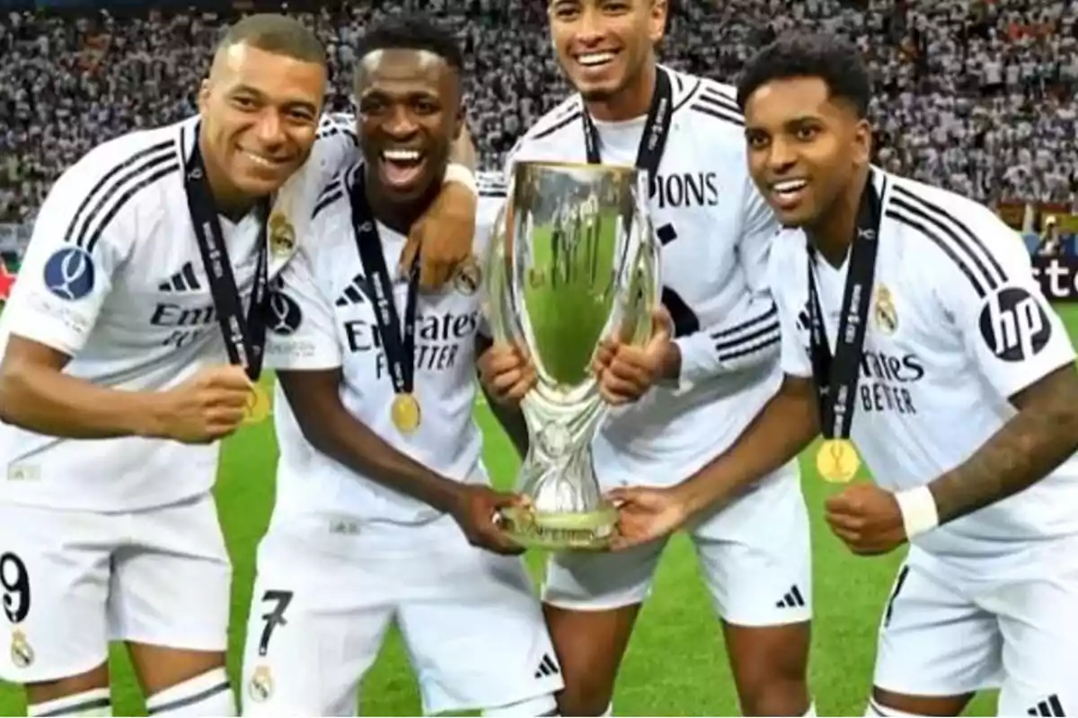 Jugadores de fútbol del Real Madrid celebran con un trofeo en el campo, vistiendo uniformes blancos y medallas.