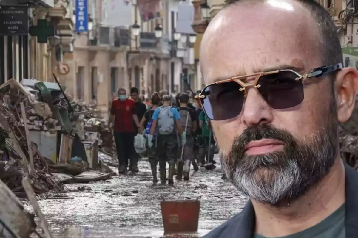 Un hombre con gafas de sol en primer plano y al fondo una calle llena de escombros y personas caminando.