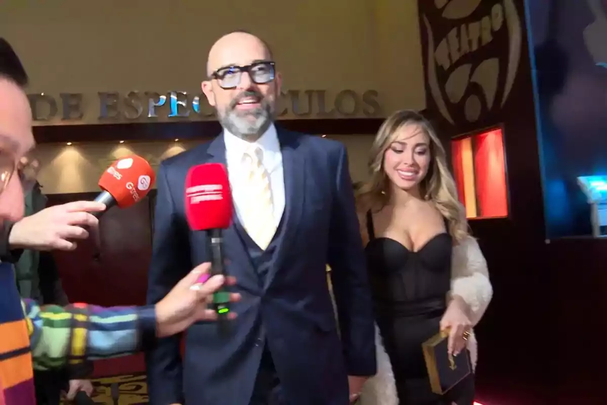 Un hombre con traje y una mujer con vestido elegante son entrevistados por reporteros en un evento.