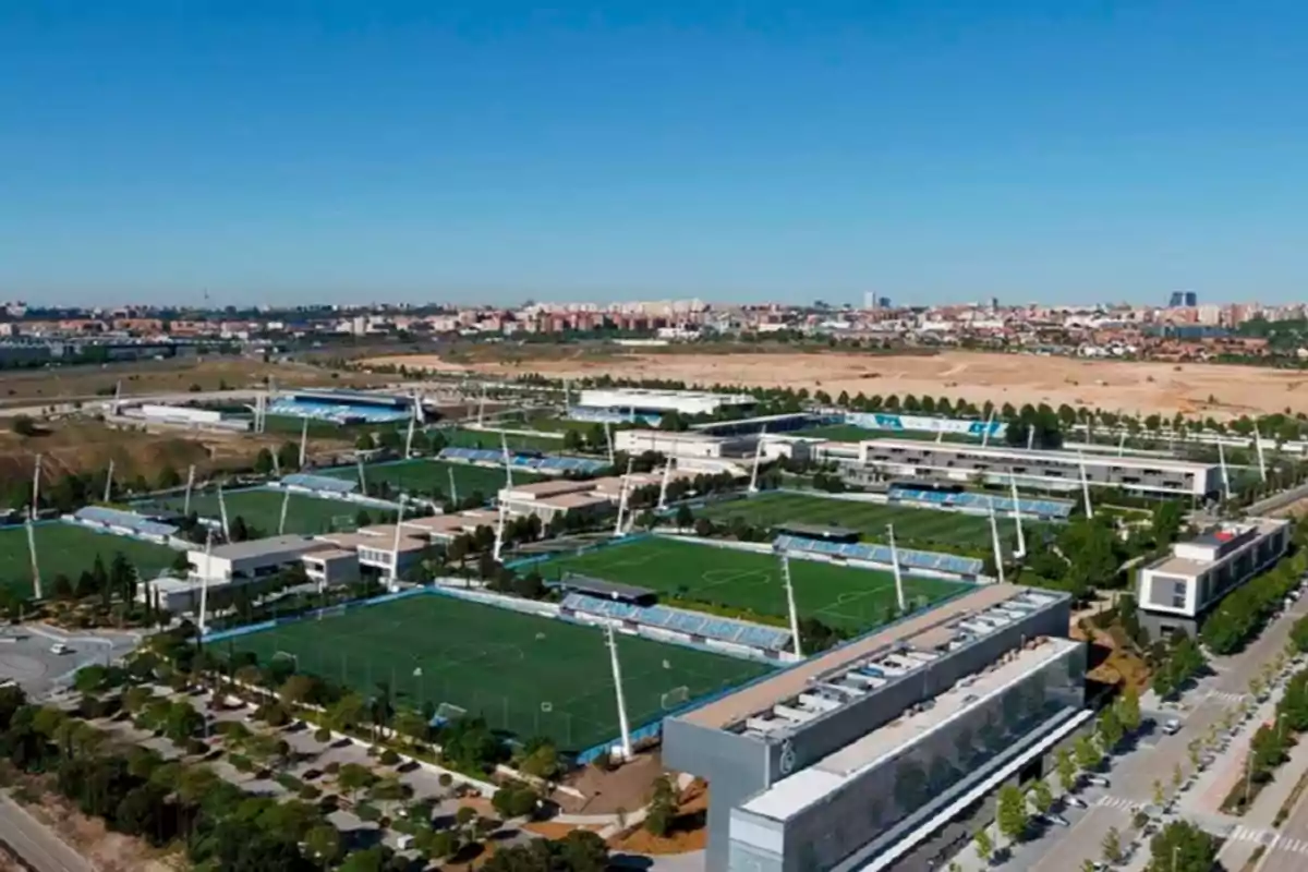 Vista aérea de un complejo deportivo con múltiples campos de fútbol y edificios administrativos en un entorno urbano.