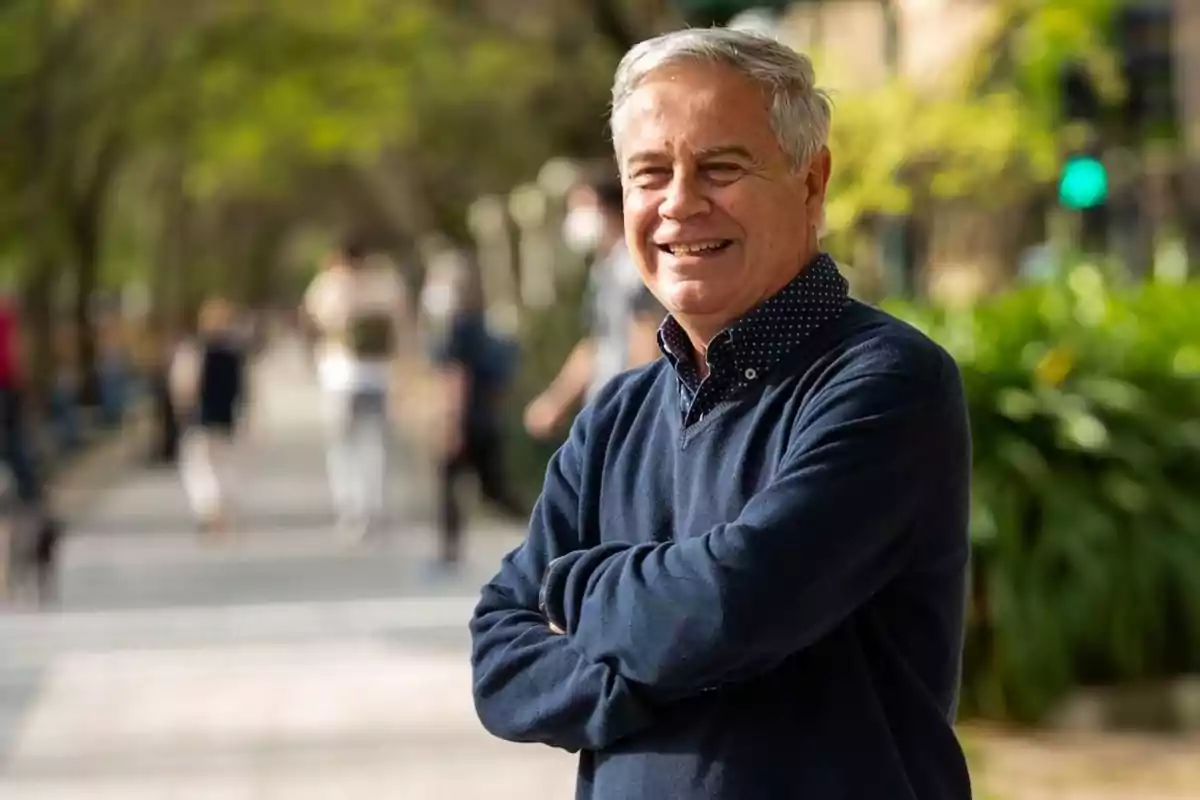 Hombre mayor sonriendo con los brazos cruzados en un parque con árboles y personas desenfocadas al fondo. Hombre mayor sonriendo con los brazos cruzados en un parque con árboles y personas desenfocadas al fondo.