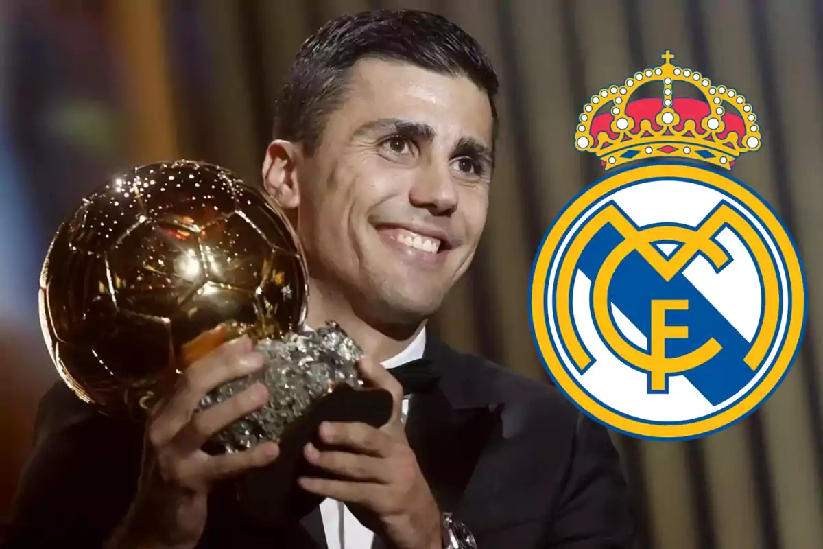 Un hombre sonriente sostiene un trofeo dorado con el logo del Real Madrid al lado.